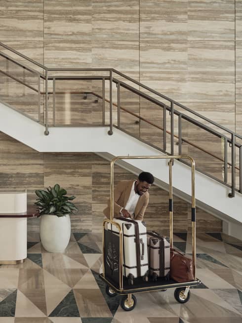 Two hotel employees wearing brown blazers and white button down shirts work in a lobby, one behind the front desk and the other loading a luggage cart