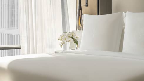 Hotel room with white bed and two benches at foot, facing blue chair, off-white wall by curtained window
