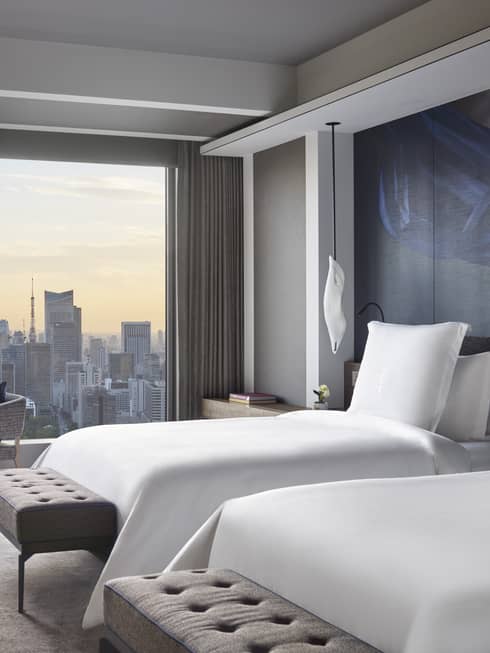 Guest room with two beds, tufted bench, seating area and floor-to-ceiling windows offering panoramic city views