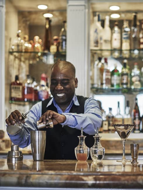 Smiling bartender pours liquor into martini cocktail shaker at bar