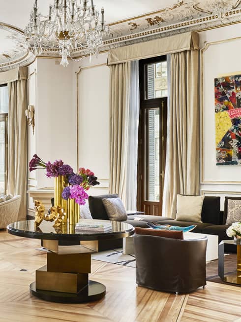 Royal suite living room with high ceilings, ornate chandeliers, grand mirror over a marble fireplace, luxury seating and modern dining table with gold accents