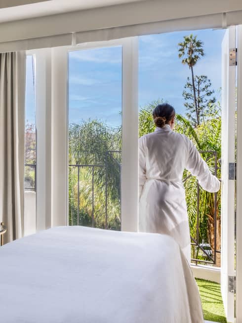 A woman walking outside from a massage room with large windowed doors and plants on one wall.