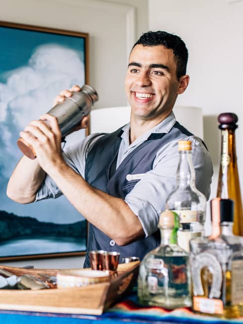 A bartender shaking a cocktail with liquor bottles on a cart in front of him.
