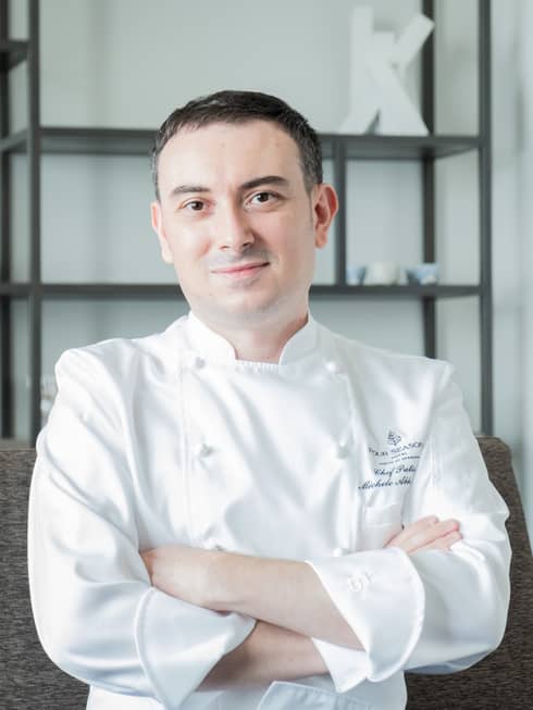 Smiling man wearing white chef's coat and sitting on couch poses for camera with arms crossed