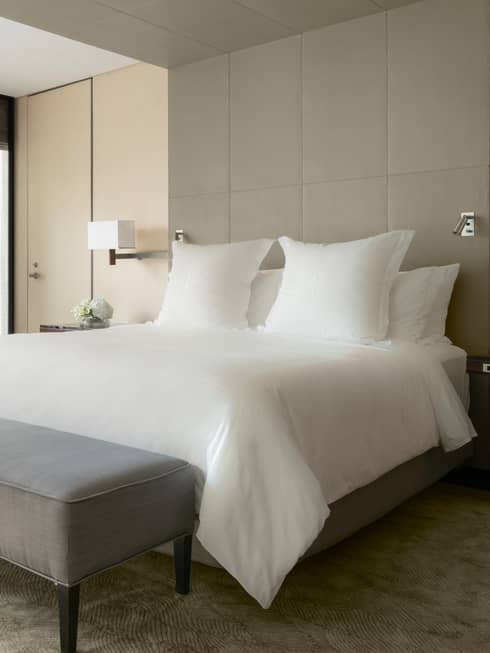 Sunny Deluxe One Bedroom Suite hotel bed with white linens, grey bench at foot