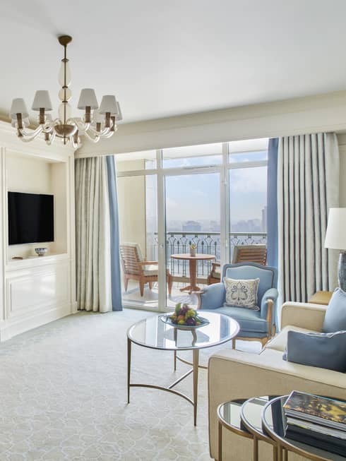 Premium Executive Suite living area with cream couch and blue accent pillows, blue armchair, sliding glass door to balcony, doorway to bedroom