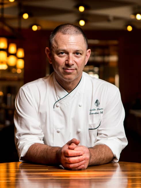 Portrait photo of Executive Chef Cyrille Pannier wearing white uniform, sitting at wood table with hands clasped in front