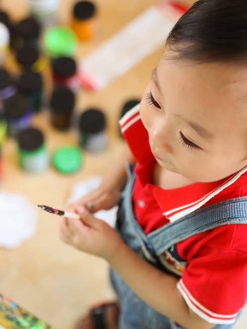 Aerial view of toddler boy standing in front of a canvas holding a paintbrush, a young girl sitting in the background surrounded by tins of paint
