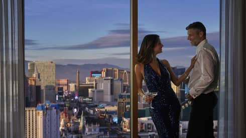 Couple holds cocktails, leans against floor to ceiling windows overlooking Las Vegas