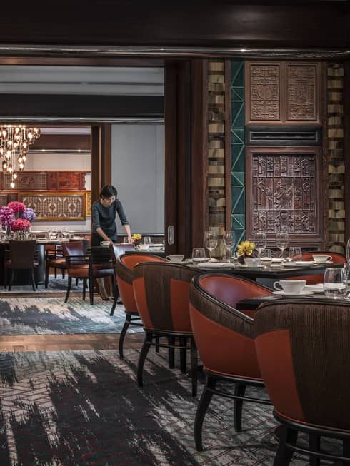 Woman sets table at Jiang-Nan Chun, Michelin-starred Cantonese restaurant, with red leather bucket chairs around dining tables, carved wood walls.