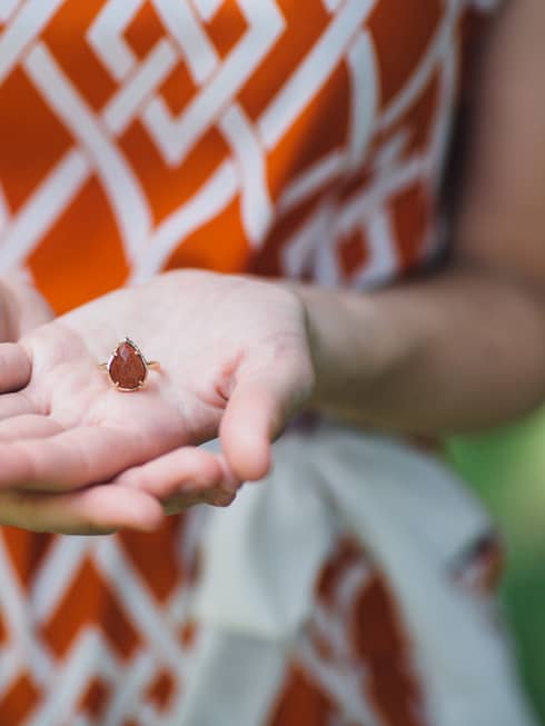 A woman displays a delicate ring with a vibrant, orange gemstone at its center