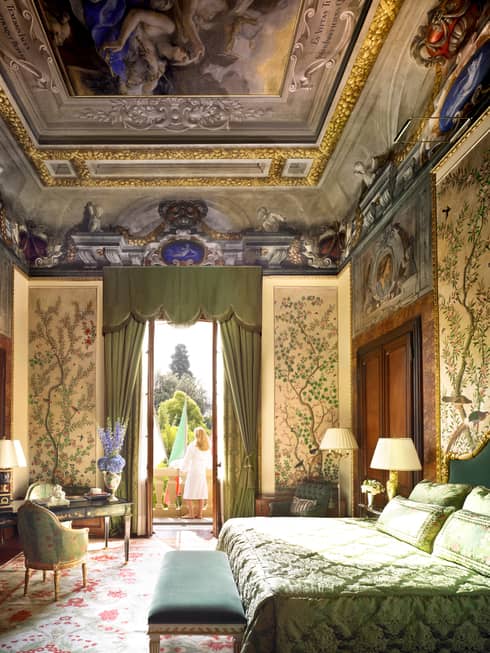 Woman in white bathrobe stands on sunny balcony by open door to bedroom, vaulted ceilings with mural