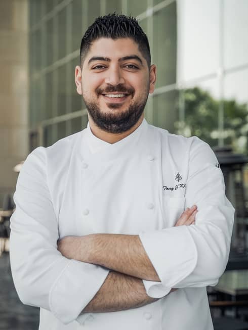 A chef in a white uniform stands with his arms crossed in front of the property.