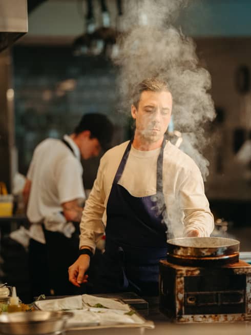 Chef wearing white long-sleeve shirt and dark-blue apron tends to a pot with rising steam