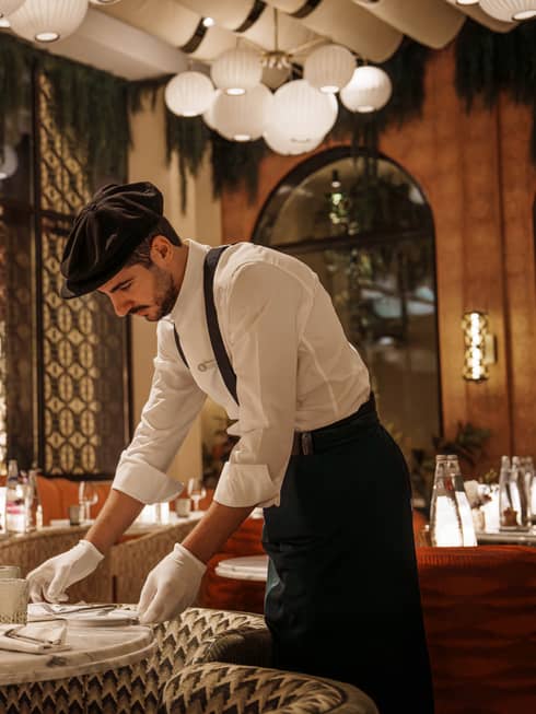 Waiter wearing white button-down shirt, black pants, black suspenders, black beret and white gloves arranges a plate on a table in an Art Deco?style restaurant