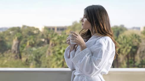 A woman in a white robe sips coffee while looking over a balcony, a variety of food on a table in front of her