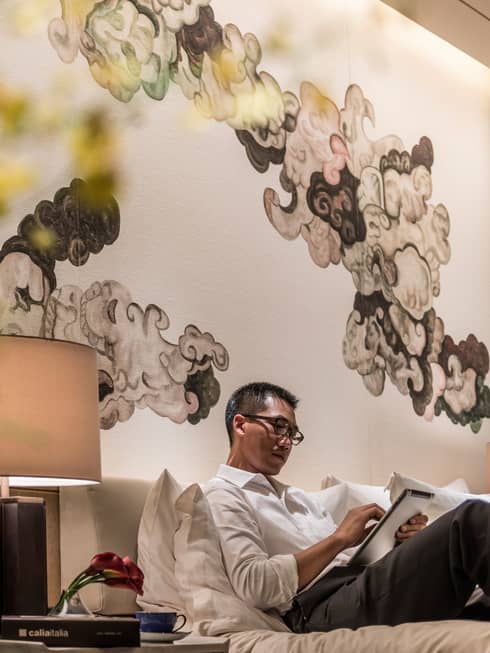 Man lounge on bed, reads iPad under black-and-white Chinese brushwork mural