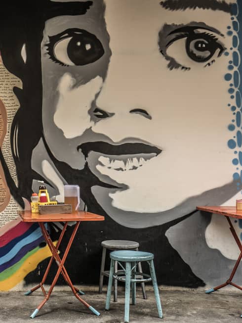 Street art mural featuring a large black-and-white face on a wall, with small, colourful dining tables and stools in front it