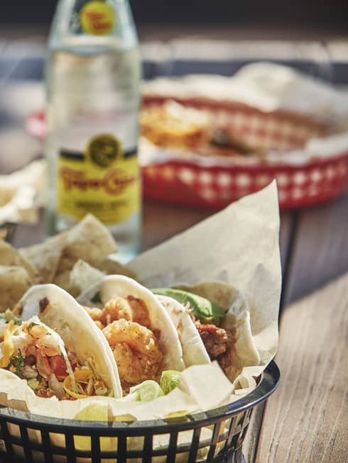 Three tacos with shrimp, shredded cheese, lettuce and tomato are arranged in a basket