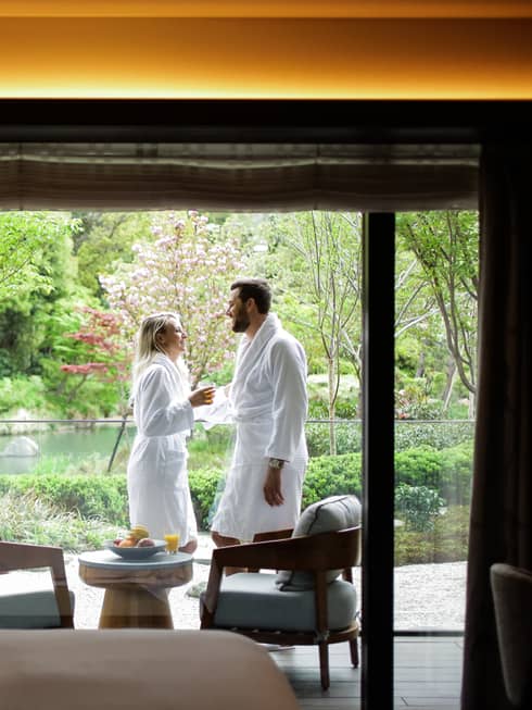 Smiling couple wearing white bathrobes on sunny balcony past hotel room bed