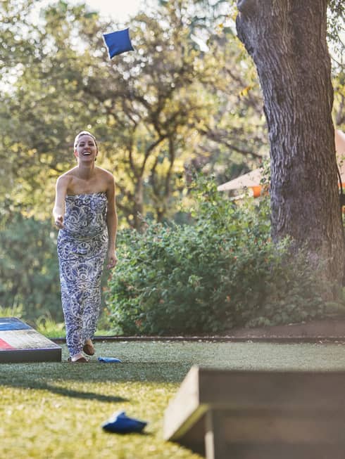 Laughing woman tosses bean bag on lawn, man holding beer watches