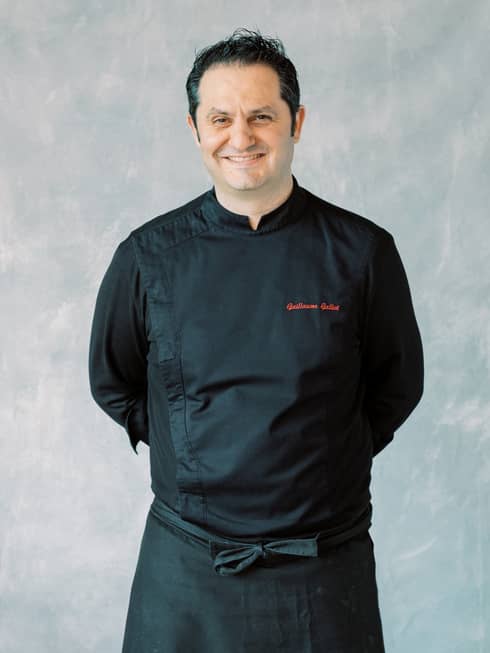 Chef Guillaume Galliot in a black chef's uniform with red embroidery, standing against a light wall. Hands behind his back, he smiles warmly at the camera.