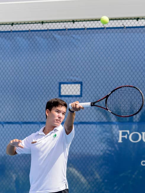 Man wearing white shirt swings racket by Tennis Centre net