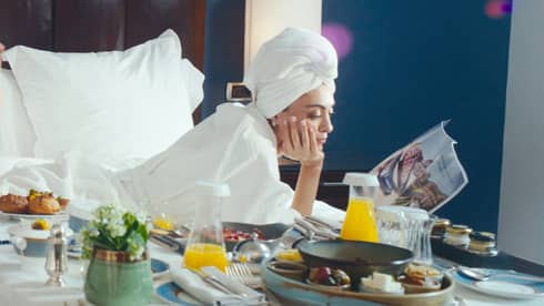 Woman relaxing and reading magazine while in a luxury hotel bed with food nearby
