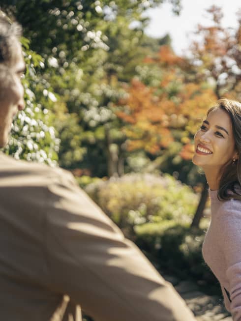 Smiling couple walks along the tree-lined pathway, pond and fall-hued trees in background