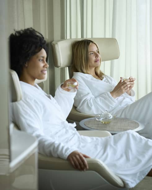 Two spa guests in white robes sitting on lounge chairs drinking champagne