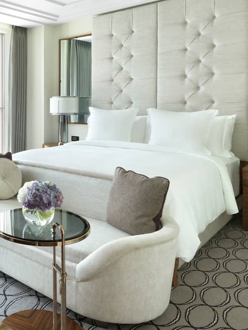 Deluxe room plush white loveseat at foot of bed by sunny window with grey curtains, table and chair