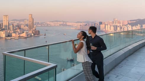 Couple stand at glass balcony, look out over city skyline, waterfront at sunset
