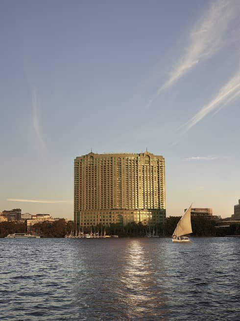 A large hotel building in the distance with a boat sailing on a river in front of it.