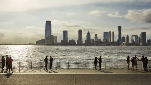 Tourists silhouetted by the setting sun on a fenced walkway by the water’s edge. Across the water are modern skyscrapers.