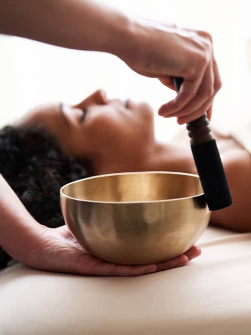 Woman with eyes closed lies on massage table in as vibrational sound therapy is performed with a Tibetan singing bowl