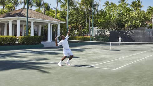 A man in all white trows a tennis ball high into the air, mid-serve 