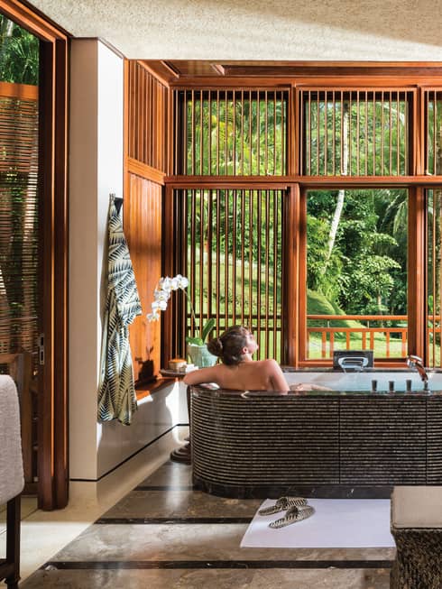 Woman with bare back relaxes in black freestanding spa tub near double sink, wood shutters open to lush garden view