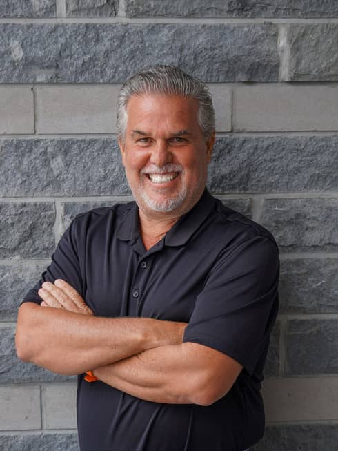 A man in a black shirt standing with his arms folded in front of a brick wall