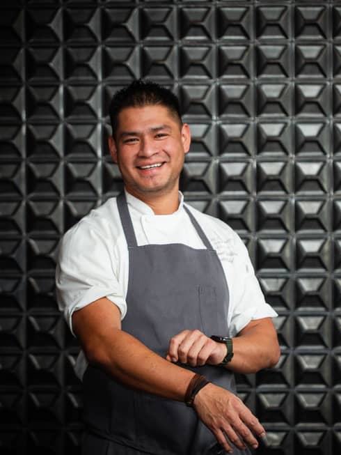 Smiling Chef Manu Calderón wearing a white shirt and grey apron against a steel background