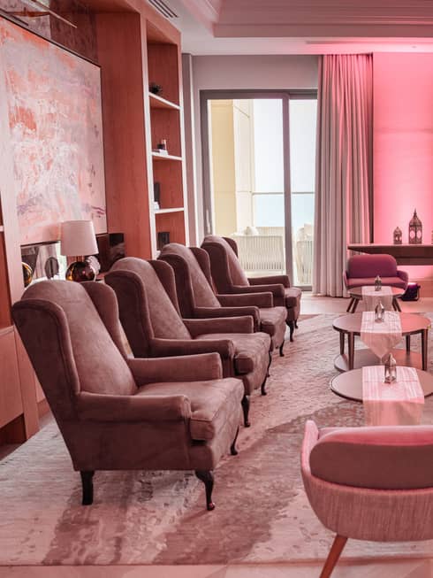 Two rows of four armchairs each face each other in a lounge with two additonal chairs on either end and three small round tables in the middle, all lit by natural light from large nearby windows