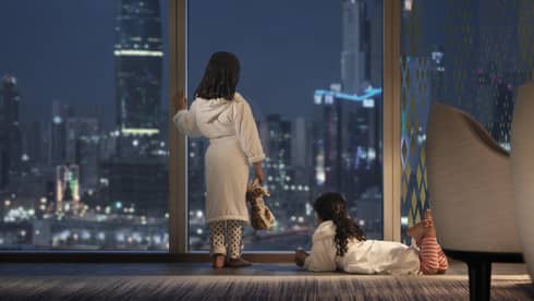 Two young children wearing bath robes look out a floor-to-ceiling window at a nighttime view of an urban cityscape.