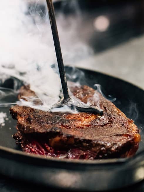 A close up of a Longhorn-Branded Steak steaming in a cast iron skillet on a silver, metal industrial counter