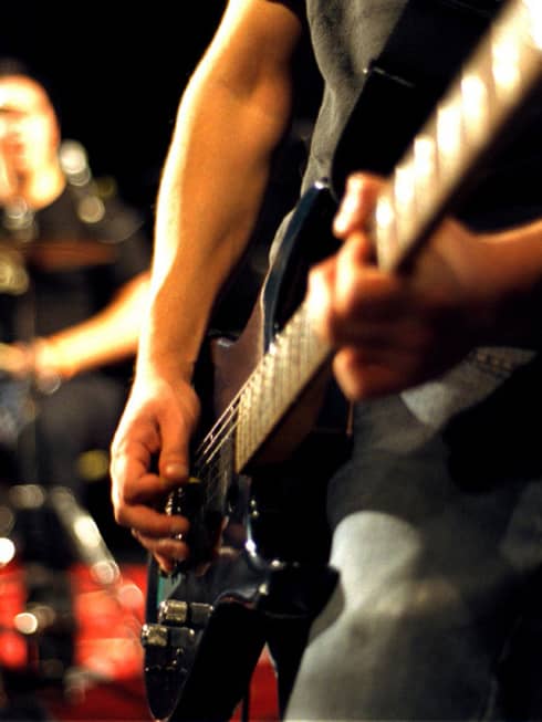 Close-up of musician's arms and hands playing an electric guitar with drumset in the background.