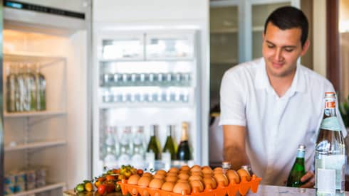 Smiling man holds green bottle in front of open fridge, fresh eggs on counter