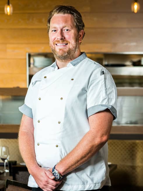 Portrait photo of smiling Executive Chef Drew Adams standing in white uniform