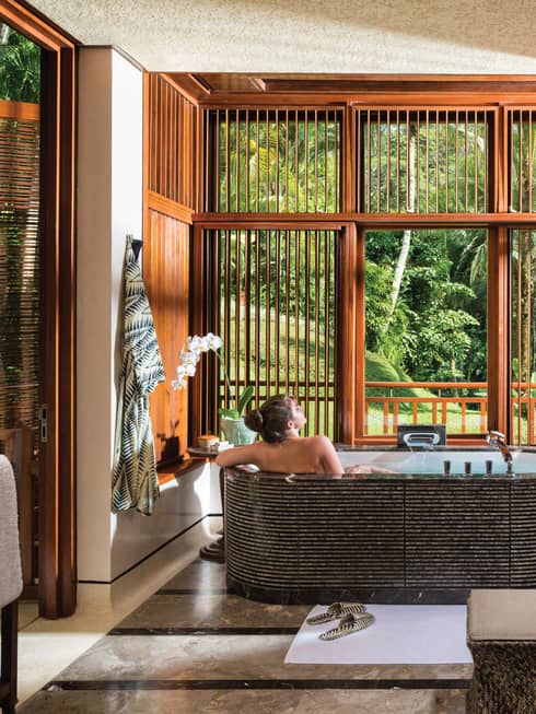 Woman with bare back relaxes in black freestanding spa tub near double sink, wood shutters open to lush garden view