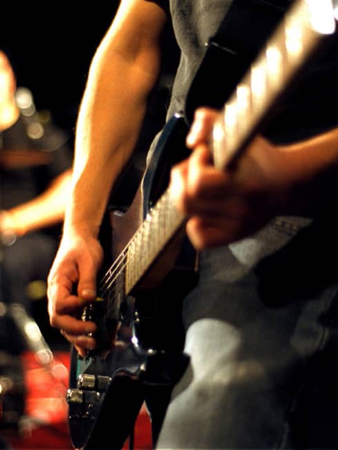 Close-up of musician's arms and hands playing an electric guitar with drumset in the background.
