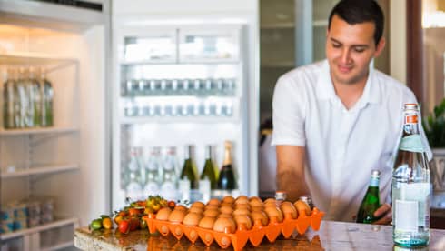 Smiling man holds green bottle in front of open fridge, fresh eggs on counter