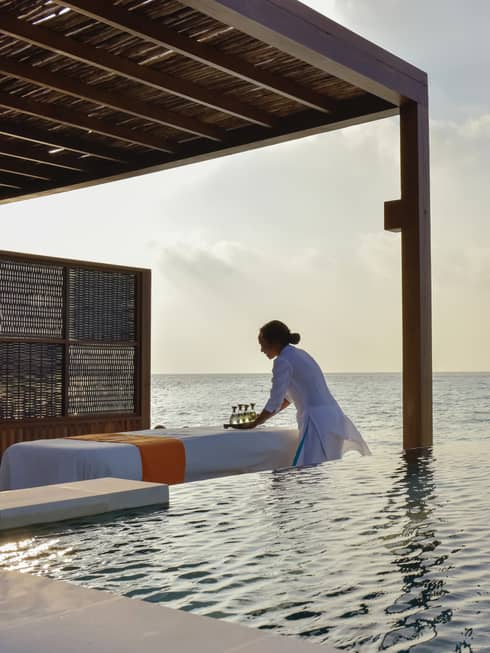 Woman sets tray down on table in front of infinity pool alongside ocean