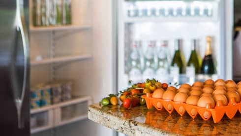 Smiling man holds green bottle in front of open fridge, fresh eggs on counter
